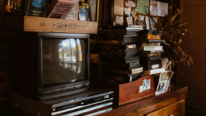 A vintage television and VCR sit in sync on a wooden cabinet next to a stack of VHS tapes and assorted books and papers.
