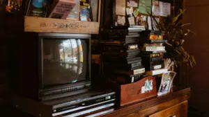 A vintage television and VCR sit in sync on a wooden cabinet next to a stack of VHS tapes and assorted books and papers.