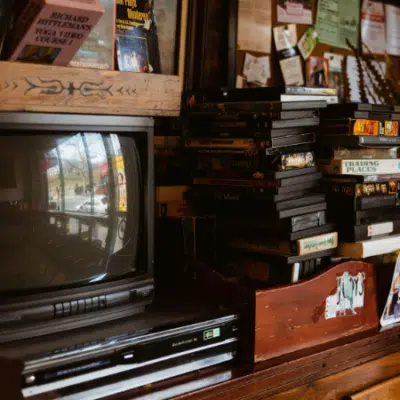 A vintage television and VCR sit in sync on a wooden cabinet next to a stack of VHS tapes and assorted books and papers.