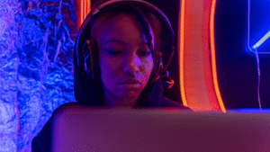 A person wearing headphones sits in front of a laptop, illuminated by blue and orange neon lights, immersed in their work as if performing for an unseen audience.