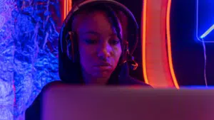 A person wearing headphones sits in front of a laptop, illuminated by blue and orange neon lights, immersed in their work as if performing for an unseen audience.