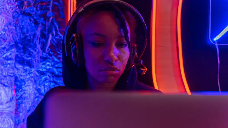 A person wearing headphones sits in front of a laptop, illuminated by blue and orange neon lights, immersed in their work as if performing for an unseen audience.