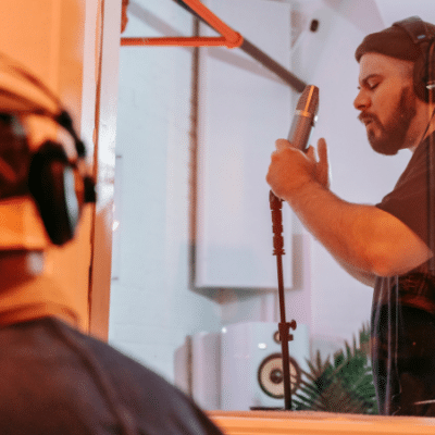 A man wearing headphones sings lyrics into a microphone in a recording studio, while another person observes from behind glass.