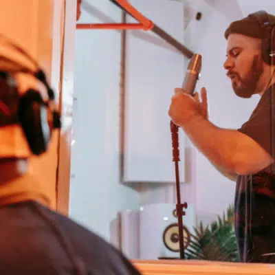 A man wearing headphones sings lyrics into a microphone in a recording studio, while another person observes from behind glass.