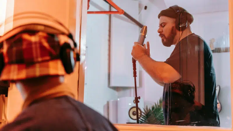 A man wearing headphones sings lyrics into a microphone in a recording studio, while another person observes from behind glass.