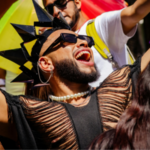 Person wearing a black spiked headpiece, pearl necklace, and ripped shirt celebrates with arms raised at an outdoor LGBTQ event, surrounded by other people and rainbow flags.