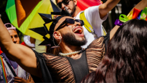 Person wearing a black spiked headpiece, pearl necklace, and ripped shirt celebrates with arms raised at an outdoor LGBTQ event, surrounded by other people and rainbow flags.