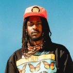An artist with long dreadlocks wears a red cap, a patterned bandana, and a black shirt with butterfly graphics and the words "Be Yourself" against a clear blue sky.