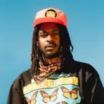 An artist with long dreadlocks wears a red cap, a patterned bandana, and a black shirt with butterfly graphics and the words "Be Yourself" against a clear blue sky.