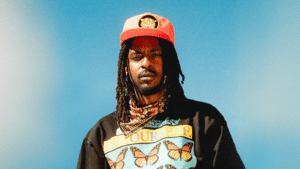 An artist with long dreadlocks wears a red cap, a patterned bandana, and a black shirt with butterfly graphics and the words "Be Yourself" against a clear blue sky.