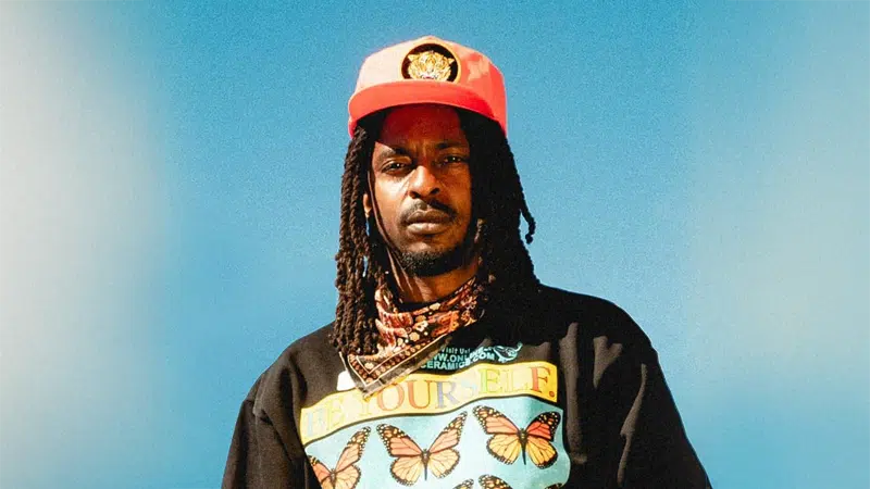 An artist with long dreadlocks wears a red cap, a patterned bandana, and a black shirt with butterfly graphics and the words "Be Yourself" against a clear blue sky.