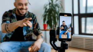 Everything You Need To Know About TikTok for Artists A man sits indoors, smiling and pointing at a smartphone on a tripod, which is filming him. A potted plant and red cup are in the background.