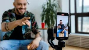 A man sits indoors, smiling and pointing at a smartphone on a tripod, which is filming him. A potted plant and red cup are in the background.