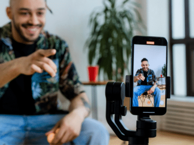 A man sits indoors, smiling and pointing at a smartphone on a tripod, which is filming him. A potted plant and red cup are in the background.