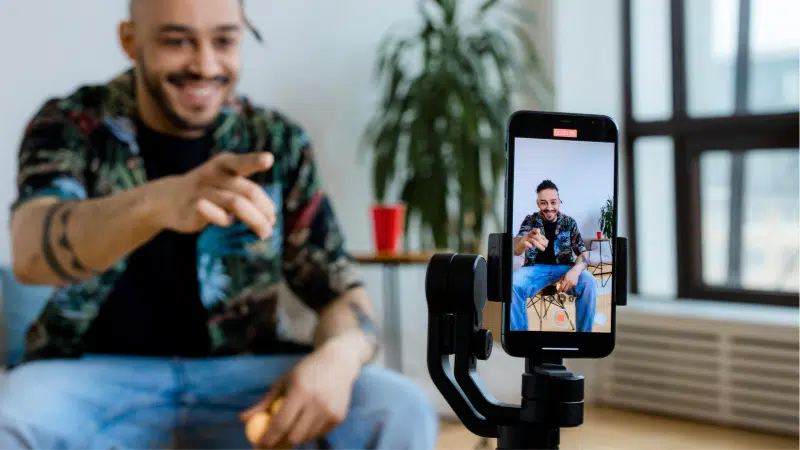 A man sits indoors, smiling and pointing at a smartphone on a tripod, which is filming him. A potted plant and red cup are in the background.