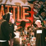 A rap battle takes place in front of a graffiti wall with a crowd watching; participants wear casual streetwear and sunglasses.