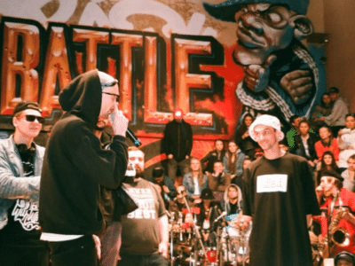 A rap battle takes place in front of a graffiti wall with a crowd watching; participants wear casual streetwear and sunglasses.