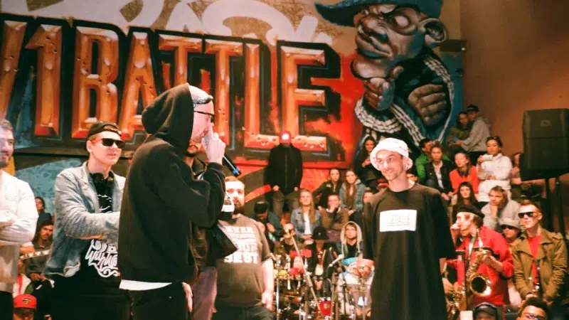 A rap battle takes place in front of a graffiti wall with a crowd watching; participants wear casual streetwear and sunglasses.