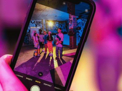 A smartphone screen displays three people posing for a photo in a colorful, graffiti-covered space under vibrant lighting, capturing a moment of joy and connection that celebrates friendship and mental health.