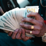 A person wearing gold jewelry, perhaps flaunting their royalties, holds and fans out a stack of U.S. dollar bills—mainly $20s and $100s—inside a car.