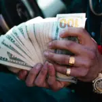 A person wearing gold jewelry, perhaps flaunting their royalties, holds and fans out a stack of U.S. dollar bills—mainly $20s and $100s—inside a car.