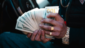 A person wearing gold jewelry, perhaps flaunting their royalties, holds and fans out a stack of U.S. dollar bills—mainly $20s and $100s—inside a car.