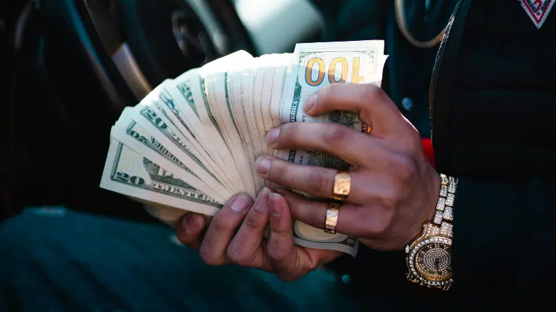 A person wearing gold jewelry, perhaps flaunting their royalties, holds and fans out a stack of U.S. dollar bills—mainly $20s and $100s—inside a car.