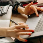 Person with red nail polish uses a laptop and writes in a notebook, with an auto calculator placed nearby on the desk.
