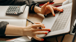 Person with red nail polish uses a laptop and writes in a notebook, with an auto calculator placed nearby on the desk.
