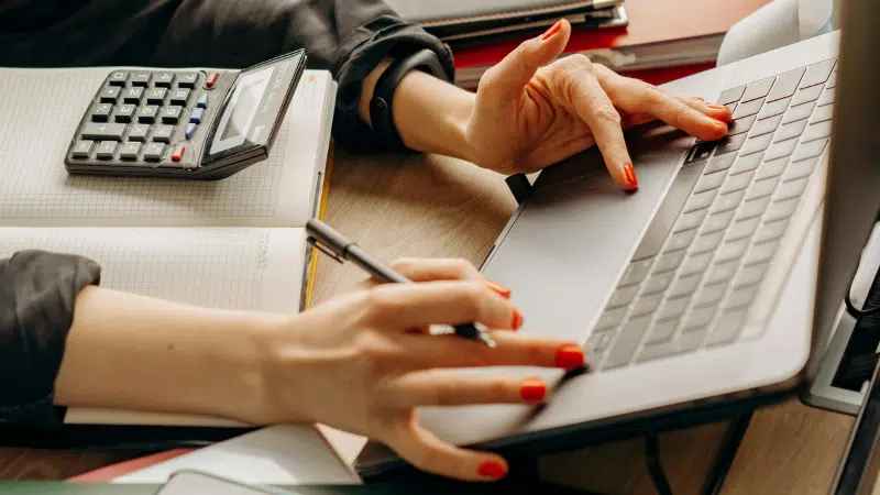 Person with red nail polish uses a laptop and writes in a notebook, with an auto calculator placed nearby on the desk.