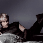 A person with short, light blonde hair is lying on a bed with white pillows, wearing a sleeveless dark shirt, long black gloves, and black pants against a plain background.