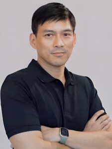 A man with short dark hair wearing a black polo shirt and a smartwatch stands with arms crossed against a plain background.
