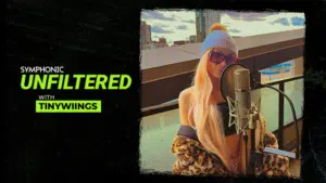 A woman with long blonde hair and sunglasses sings into a microphone on a rooftop. Text reads: "Symphonic Unfiltered with Tinywiings.