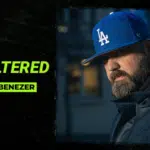 A man in a blue LA baseball cap and dark jacket stands outside at dusk; text reads "Symphonic Unfiltered with Evil Ebenezer.