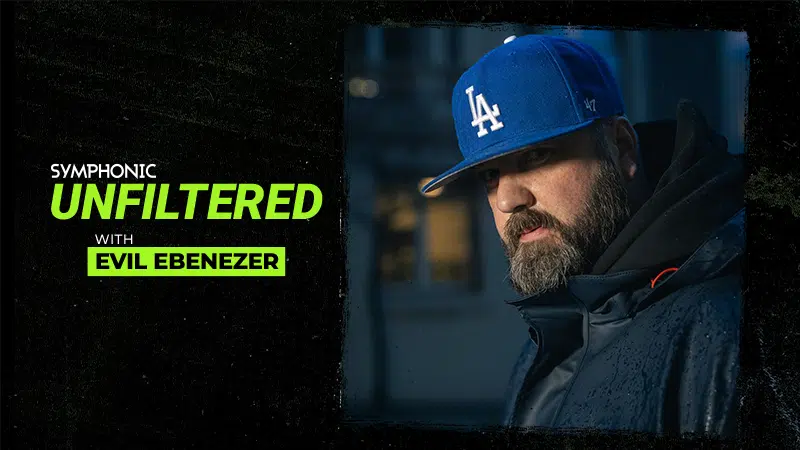 A man in a blue LA baseball cap and dark jacket stands outside at dusk; text reads "Symphonic Unfiltered with Evil Ebenezer.
