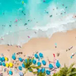 Aerial view of a crowded beach with people swimming in turquoise water, relaxing under blue umbrellas on the sand, and enjoying a lively playlist drifting through the summer air.