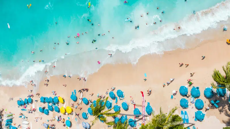 Aerial view of a crowded beach with people swimming in turquoise water, relaxing under blue umbrellas on the sand, and enjoying a lively playlist drifting through the summer air.