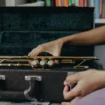 A person’s hand places a brass trumpet into a black, velvet-lined case—perhaps after a scholarship audition—with a bookshelf visible in the background.