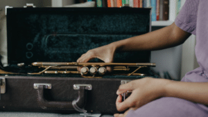 A person’s hand places a brass trumpet into a black, velvet-lined case—perhaps after a scholarship audition—with a bookshelf visible in the background.
