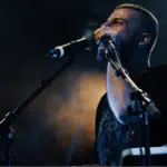 A man stands at a microphone on a dimly lit stage, singing or speaking as he prepares for the release, with a keyboard and additional microphone visible in front of him.