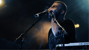 What Makes a Smart Music Release Strategy in 2025? A man stands at a microphone on a dimly lit stage, singing or speaking as he prepares for the release, with a keyboard and additional microphone visible in front of him.