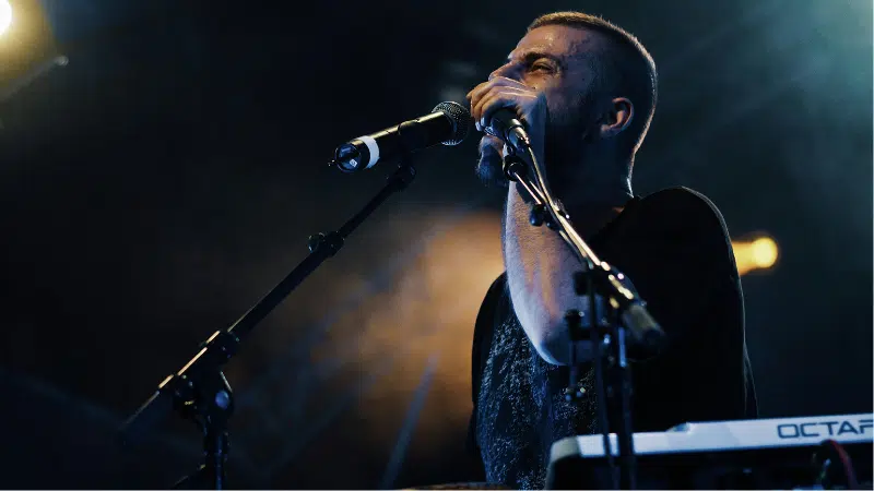 A man stands at a microphone on a dimly lit stage, singing or speaking as he prepares for the release, with a keyboard and additional microphone visible in front of him.