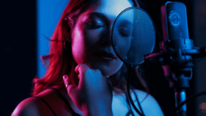 A woman stands in front of a microphone with a pop filter, singing or recording music in a dimly lit studio bathed in blue and pink lighting.