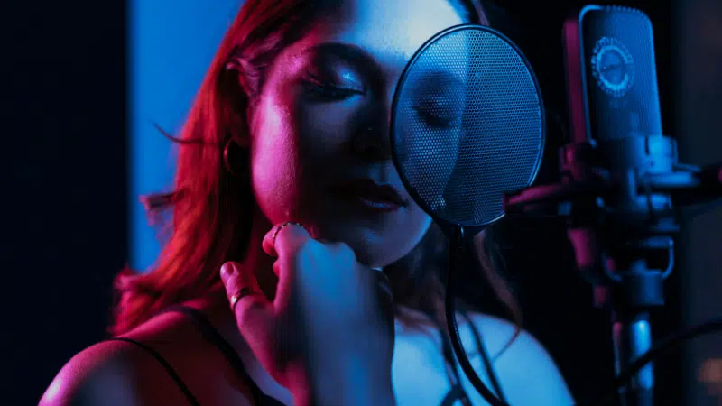 A woman stands in front of a microphone with a pop filter, singing or recording music in a dimly lit studio bathed in blue and pink lighting.