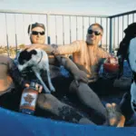 Three men with tattoos relax in a small inflatable pool with drinks on a sunny summer day, joined by two dogs, including a Dalmatian.