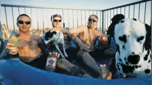 Three men with tattoos relax in a small inflatable pool with drinks on a sunny summer day, joined by two dogs, including a Dalmatian.