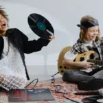 Two young musicians sit on a rug with music equipment; one holds a vinyl record and sings, while the other plays an electric guitar. Loose records and a microphone are scattered on the floor.