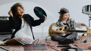 Two young musicians sit on a rug with music equipment; one holds a vinyl record and sings, while the other plays an electric guitar. Loose records and a microphone are scattered on the floor.