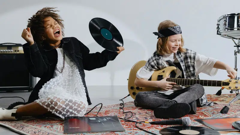 Two young musicians sit on a rug with music equipment; one holds a vinyl record and sings, while the other plays an electric guitar. Loose records and a microphone are scattered on the floor.