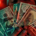 A person holds up several spread-out $100 bills, fanned in front of their face, with a warm red glow in the background—illustrating potential benefits from the HITS Act.